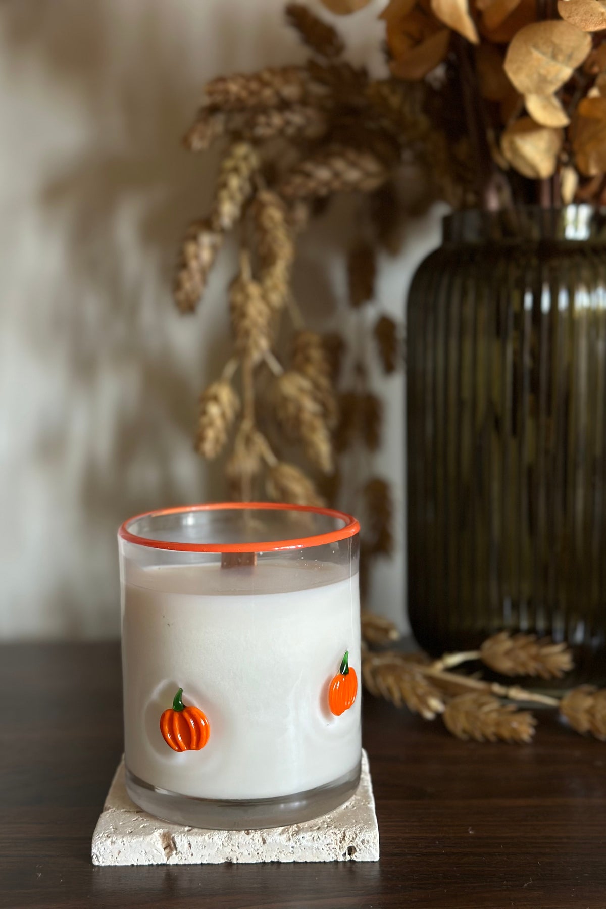 Candle with pumpkin decorations on a wooden surface with a vase and dried plants in the background.