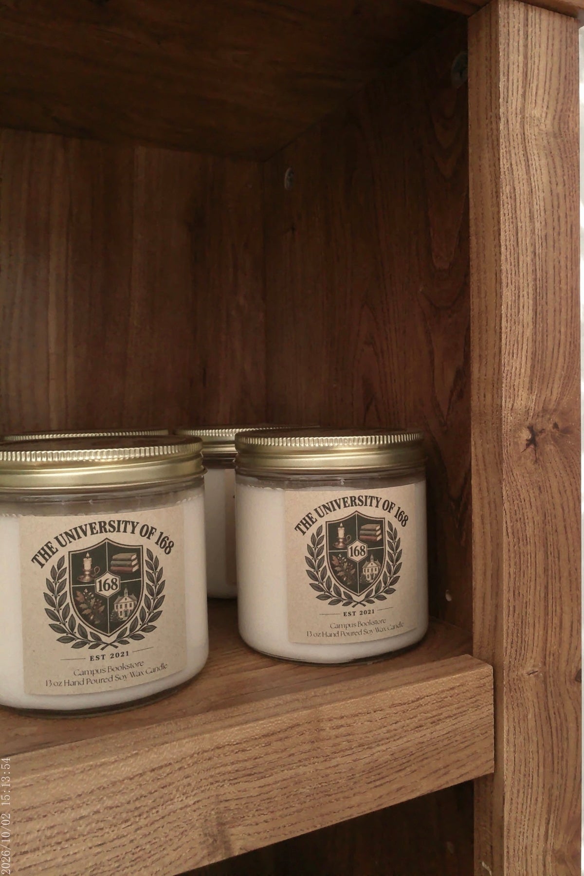 Three white candle jars with gold lids and a black crest label reading 'THE UNIVERSITY OF 186' displayed on a wooden shelf.